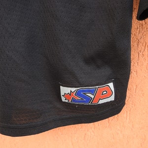 Pode incluir: Close-up de uma camisa de malha preta. A camisa tem uma etiqueta com as letras "SP" em azul e vermelho, com um design de folha de bordo. A camisa &eacute; feita de um tecido respir&aacute;vel e texturizado.