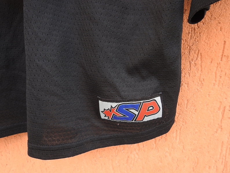Pode incluir: Close-up de uma camisa de malha preta. A camisa tem uma etiqueta com as letras "SP" em azul e vermelho, com um design de folha de bordo. A camisa &eacute; feita de um tecido respir&aacute;vel e texturizado.
