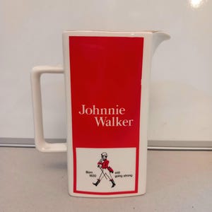 May include: A white ceramic pitcher with a rectangular handle and a spout. The front features a red rectangle with "Johnnie Walker" in white text. Below, a white rectangle shows a walking figure and the text "Born 1820 still going strong."