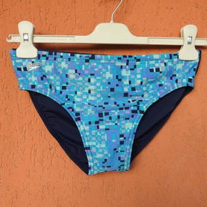 May include: A pair of swim briefs featuring a blue, light blue, and dark blue pixelated digital camouflage pattern. The garment has a solid dark blue interior lining and displays the Speedo brand logo in white on the upper left side.