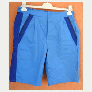 May include: Light blue shorts with dark blue side panels and trim, hanging on a white hanger. The shorts have a pleated front and a tailored design. The fabric appears to be a lightweight material, suitable for warm weather.