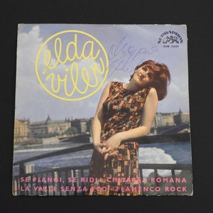 Puede incluir: Un disco de vinilo de 45 rpm vintage de Elda Viler, con las canciones "Se Plangi, Se Rid" y "Chitarra Romana". La portada del disco presenta una foto de Elda Viler con un vestido estampado, de pie en un balcón con vistas a una ciudad.