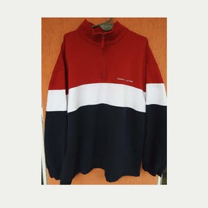 Puede incluir: Una sudadera con bloques de color en rojo, blanco y azul marino. La sección superior es roja, seguida de una franja blanca y una parte inferior azul marino. Tiene un cuello alto con cierre de cremallera y el texto "TOMMY HILFIGER" en blanco.