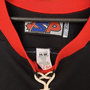 Pode incluir: Camisola de h&oacute;quei preta com acabamento vermelho ao redor da gola e das mangas. A camisola tem um decote com cadar&ccedil;o branco e uma etiqueta com o texto "H.W. MADE IN/FAIT AU HECHO EN CANADA". A camisola tamb&eacute;m tem um logotipo com as letras "SP".