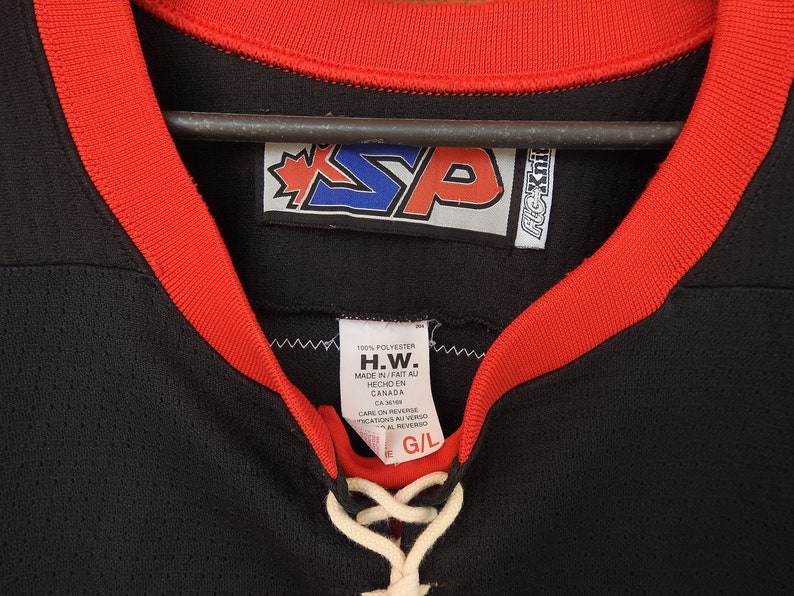 Pode incluir: Camisola de h&oacute;quei preta com acabamento vermelho ao redor da gola e das mangas. A camisola tem um decote com cadar&ccedil;o branco e uma etiqueta com o texto "H.W. MADE IN/FAIT AU HECHO EN CANADA". A camisola tamb&eacute;m tem um logotipo com as letras "SP".