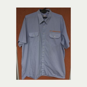 Puede incluir: Camisa de manga corta a rayas azules y blancas con dos bolsillos en el pecho. La camisa tiene el texto "Lufthansa Technik" bordado en naranja en el pecho derecho. La camisa está colgada en una percha negra.