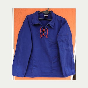 May include: A blue work shirt with long sleeves, a collar, and two front pockets. The shirt features a red lace-up detail at the neckline. The garment is hanging on a black hanger against an orange background.