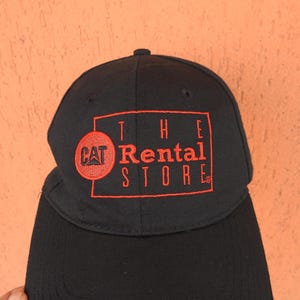 May include: A black baseball cap featuring an embroidered red rectangular border and the text "THE Rental STORE" in red. To the left of the text, a red circular patch contains the black "CAT" logo. The cap has a structured crown and a curved brim.