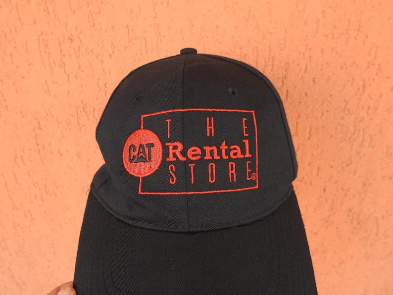 May include: A black baseball cap featuring an embroidered red rectangular border and the text "THE Rental STORE" in red. To the left of the text, a red circular patch contains the black "CAT" logo. The cap has a structured crown and a curved brim.
