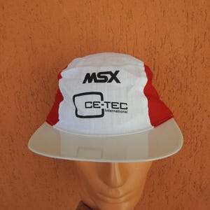 May include: White and red bucket hat with a wide brim. The hat features the text "MSX" and "CE-TEC International" in black. The hat is displayed on a mannequin head against an orange background.