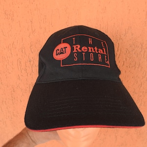 May include: A black baseball cap featuring an embroidered red logo on the front. The logo includes a red circle with the text CAT and a red rectangular outline containing the text THE RENTAL STORE. The brim has a thin red trim along the edge.