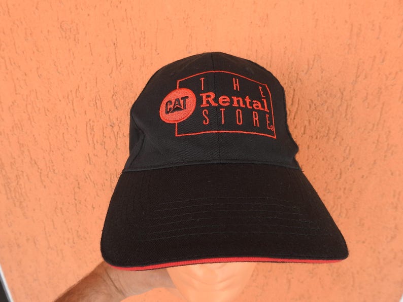 May include: A black baseball cap featuring an embroidered red logo on the front. The logo includes a red circle with the text CAT and a red rectangular outline containing the text THE RENTAL STORE. The brim has a thin red trim along the edge.