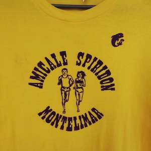 Könnte beinhalten: Ein leuchtend gelbes Kurzarm-T-Shirt mit einem dunkelvioletten Grafikdruck. Das Design zeigt den Text AMICALE SPIRIDON MONTELIMAR um eine Illustration von zwei laufenden Personen, ergänzt durch ein kleines stilisiertes Delfin-Symbol.