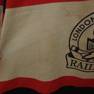 Pode incluir: Close-up de uma camisola de h&oacute;quei com corpo creme, detalhes em vermelho e preto. A camisola apresenta o texto "LONDON RAIDERS" num emblema circular com um capacete de cavaleiro e um stick de h&oacute;quei.