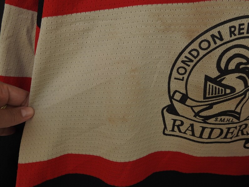 Pode incluir: Close-up de uma camisola de h&oacute;quei com corpo creme, detalhes em vermelho e preto. A camisola apresenta o texto "LONDON RAIDERS" num emblema circular com um capacete de cavaleiro e um stick de h&oacute;quei.