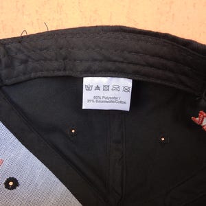May include: The interior of a black baseball cap featuring a white fabric care label. The label displays five standard laundry symbols and the text 65% Polyester / 35% Baumwolle/Cotton. The cap has black stitching and a red embroidered detail.