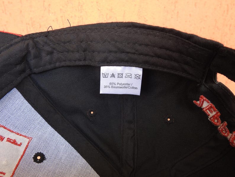 May include: The interior of a black baseball cap featuring a white fabric care label. The label displays five standard laundry symbols and the text 65% Polyester / 35% Baumwolle/Cotton. The cap has black stitching and a red embroidered detail.