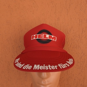 May include: A bright red trucker cap featuring the word HELM in white letters over a black circular graphic on the front panel. The curved brim displays the white printed text Wir sind die Meister fürs Auto along the outer edge of the visor.