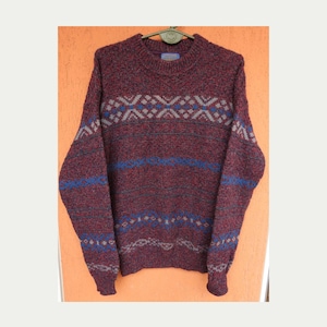 Könnte beinhalten: Ein langärmeliger Pullover mit Rundhalsausschnitt aus dunkelburgunderfarbenem, meliertem Strick. Das Kleidungsstück zeigt horizontale Bänder mit geometrischen Mustern in Blau- und Grautönen auf Brust und Ärmeln sowie gerippte Bündchen am Kragen.