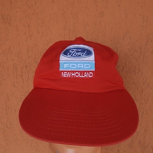 May include: A bright red baseball cap featuring an embroidered patch on the front. The patch displays the blue Ford logo, the word FORD in light blue, and the text NEW HOLLAND in white on a red background. The cap has a curved brim.