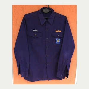 Op de afbeelding: Een donkerblauw shirt met lange mouwen, twee borstzakken en een kraag. Het shirt heeft een patch met tekst en een badge met een lelie-ontwerp. Een Duitse vlagpatch is ook zichtbaar.