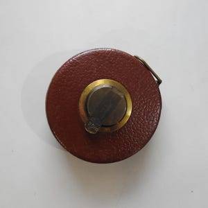 Könnte beinhalten: Ein Vintage-Maßband aus braunem Leder mit einem Messingmittelstück und einem Metallverschluss. Das Maßband ist rund und hat eine strukturierte Oberfläche. Das Messingmittelstück trägt die Gravur "2019".