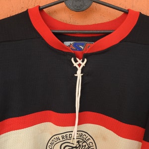 Pode incluir: Camisola de h&oacute;quei preta e vermelha com decote com atacadores brancos. A camisola apresenta uma gola vermelha e uma faixa branca e vermelha no peito. O log&oacute;tipo do London Red Circle Club &eacute; vis&iacute;vel na parte inferior.