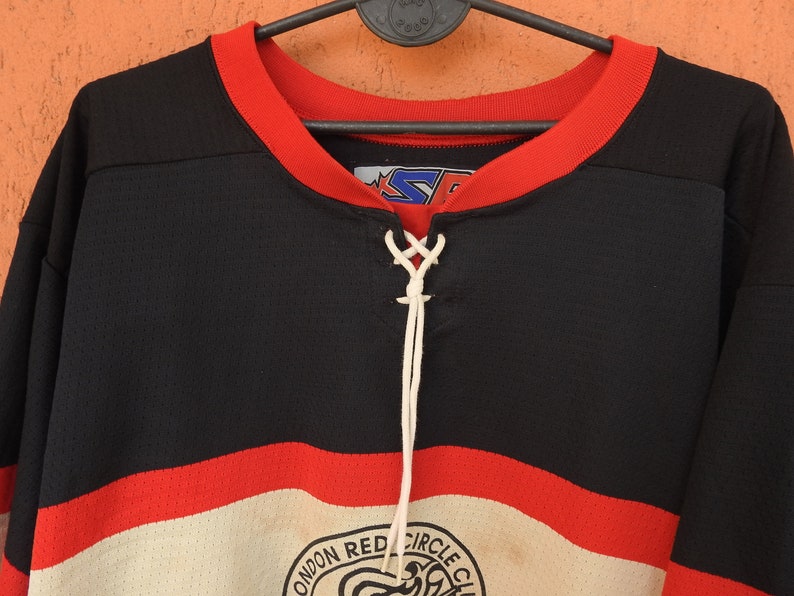 Pode incluir: Camisola de h&oacute;quei preta e vermelha com decote com atacadores brancos. A camisola apresenta uma gola vermelha e uma faixa branca e vermelha no peito. O log&oacute;tipo do London Red Circle Club &eacute; vis&iacute;vel na parte inferior.