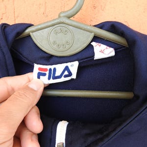 Puede incluir: Una chaqueta azul marino oscuro con cremallera, cuello blanco y una etiqueta interior blanca con el logotipo de la marca Fila en texto rojo y azul. La chaqueta se muestra en una percha de pl&aacute;stico verde con el texto MIS 2000 grabado en el centro.