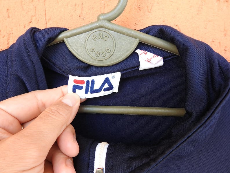 Puede incluir: Una chaqueta azul marino oscuro con cremallera, cuello blanco y una etiqueta interior blanca con el logotipo de la marca Fila en texto rojo y azul. La chaqueta se muestra en una percha de pl&aacute;stico verde con el texto MIS 2000 grabado en el centro.