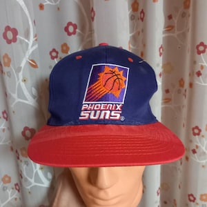 Puede incluir: Gorra de béisbol de los Phoenix Suns, azul marino y roja. La gorra presenta el logotipo del equipo, una pelota de baloncesto dentro de un diseño de sol y el nombre del equipo. La visera es roja y la corona es azul marino.