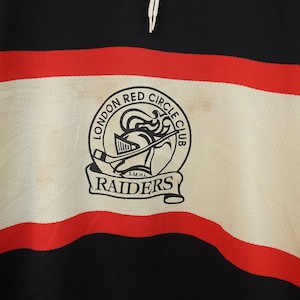 Pode incluir: Uma camisola de h&oacute;quei vintage em preto, vermelho e branco. A camisola apresenta um emblema circular com o texto "LONDON RED CIRCLE CLUB RAIDERS" e um gr&aacute;fico de capacete de cavaleiro. A camisola tem uma textura de malha.