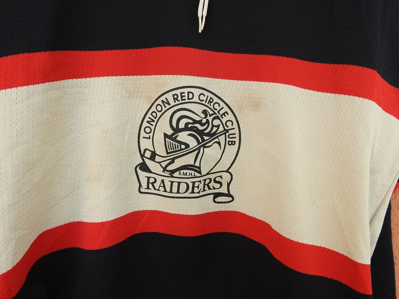 Pode incluir: Uma camisola de h&oacute;quei vintage em preto, vermelho e branco. A camisola apresenta um emblema circular com o texto "LONDON RED CIRCLE CLUB RAIDERS" e um gr&aacute;fico de capacete de cavaleiro. A camisola tem uma textura de malha.