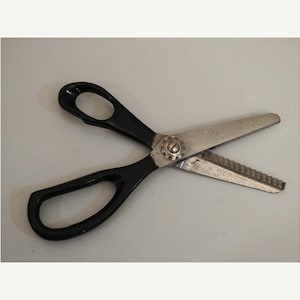 May include: A pair of metal pinking shears featuring two black plastic handles and silver-colored serrated blades. The blades are joined by a central metal screw mechanism, designed to create a zigzag pattern when cutting through various materials.