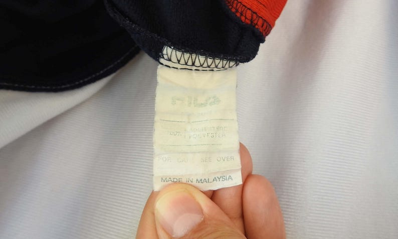 Puede incluir: Una etiqueta de cuidado rectangular blanca unida a una tela azul marino oscuro y roja. La etiqueta presenta el nombre de la marca Fila en la parte superior, seguido de texto que indica una composici&oacute;n 100% poli&eacute;ster y origen Malasia.
