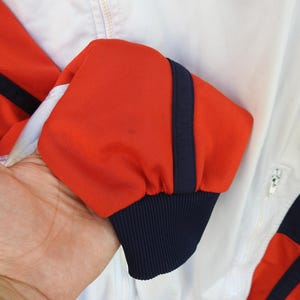 Puede incluir: Una chaqueta deportiva blanca con mangas de color naranja vibrante que presentan una franja vertical azul marino oscuro a lo largo del brazo. La manga termina en un pu&ntilde;o acanalado azul marino oscuro y la chaqueta incluye una cremallera blanca.