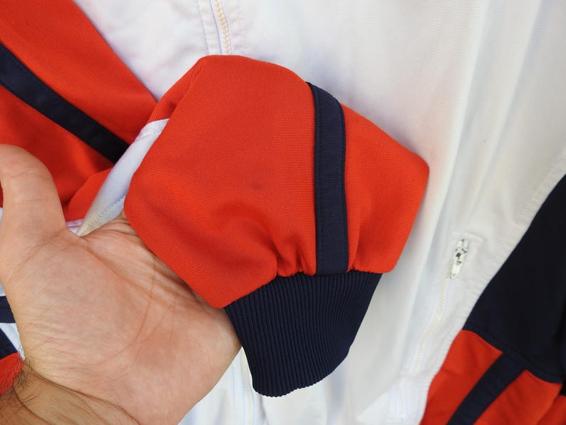 Puede incluir: Una chaqueta deportiva blanca con mangas de color naranja vibrante que presentan una franja vertical azul marino oscuro a lo largo del brazo. La manga termina en un pu&ntilde;o acanalado azul marino oscuro y la chaqueta incluye una cremallera blanca.