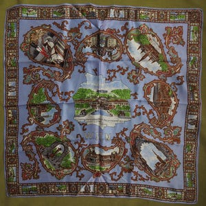 May include: A square light blue scarf featuring a central illustration of a palace with the text WIEN below it. The design includes multiple ornate paisley-shaped frames containing detailed architectural scenes of Vienna and a decorative border.