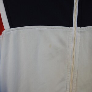 Puede incluir: Una chaqueta deportiva blanca con cremallera central completa, un panel superior negro en el pecho y un panel rojo en el lado izquierdo. La prenda incluye un peque&ntilde;o logotipo bordado en la parte inferior derecha y una mancha tenue.