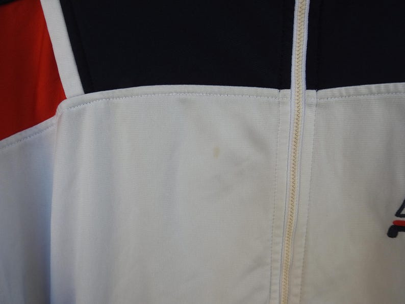 Puede incluir: Una chaqueta deportiva blanca con cremallera central completa, un panel superior negro en el pecho y un panel rojo en el lado izquierdo. La prenda incluye un peque&ntilde;o logotipo bordado en la parte inferior derecha y una mancha tenue.