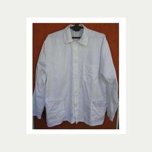 May include: A white button-down work shirt with a collar, two chest pockets, and two lower pockets. The long-sleeved shirt is made of a plain weave fabric and has a relaxed fit. The shirt is hanging on a hanger.