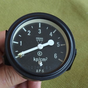Può includere: Un manometro circolare nero con un ago bianco e segni bianchi. Il manometro legge da 0 a 6 kp/cm2. Il manometro è etichettato "PREMA" e "AP-6".