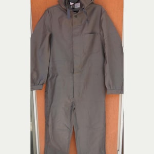 Könnte beinhalten: Grauer Kapuzen-Overall mit einer Vordertasche und Knopfverschluss. Das einteilige Arbeitskleidungsstück hat lange Ärmel und eine Kapuze mit Kordelzug. Der Overall ist aus strapazierfähigem Stoff gefertigt.