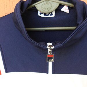 Puede incluir: Una chaqueta deportiva de color azul marino, blanco y naranja con cuello alto y cierre de cremallera frontal. El tirador de la cremallera es un dije met&aacute;lico cuadrado con el logotipo de Fila. Una etiqueta de Fila est&aacute; cosida en el cuello.