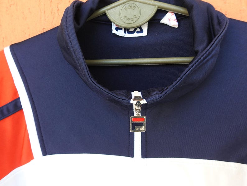 Puede incluir: Una chaqueta deportiva de color azul marino, blanco y naranja con cuello alto y cierre de cremallera frontal. El tirador de la cremallera es un dije met&aacute;lico cuadrado con el logotipo de Fila. Una etiqueta de Fila est&aacute; cosida en el cuello.