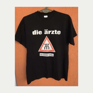 May include: A black short-sleeved t-shirt featuring the white text die ärzte above a red and white triangular road sign graphic with black symbols and the white text ES WIRD ENG. printed inside a white rectangle below the triangle.