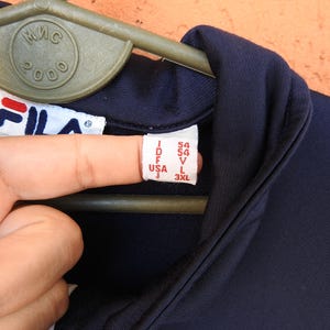 Puede incluir: Una chaqueta deportiva Fila de color azul marino oscuro con cuello alto y etiqueta de marca blanca. La etiqueta interior muestra informaci&oacute;n de tallas que incluye I 54, D 54, F 54, USA L y J 3XL. La prenda cuelga de una percha de pl&aacute;stico verde.