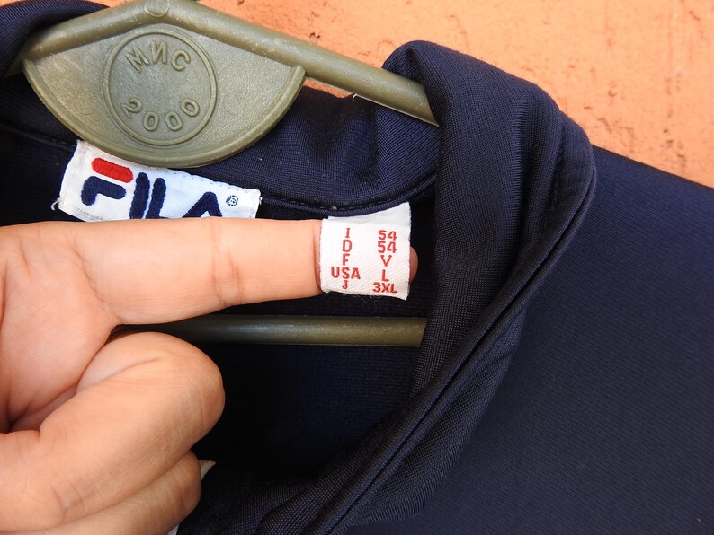 Puede incluir: Una chaqueta deportiva Fila de color azul marino oscuro con cuello alto y etiqueta de marca blanca. La etiqueta interior muestra informaci&oacute;n de tallas que incluye I 54, D 54, F 54, USA L y J 3XL. La prenda cuelga de una percha de pl&aacute;stico verde.