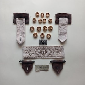 Puede incluir: Una colección de artículos de costura que incluye doce botones redondos de madera pequeños, dos lengüetas de tela de ante marrón oscuro con extensiones de tela beige claro, dos lengüetas de ante marrón oscuro con herrajes metálicos, una tira de tela beige bordada y una pequeña etiqueta metálica rectangular con el texto LANDHAUS C&A NATURALLY.