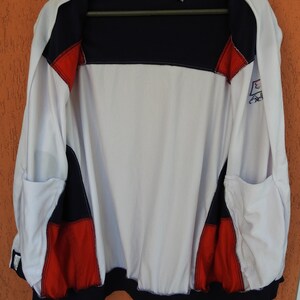 Puede incluir: Una chaqueta deportiva con cremallera Fila que presenta un dise&ntilde;o de bloques de color en blanco, azul marino y rojo. La chaqueta tiene un cuerpo principal blanco, hombros y dobladillo azul marino, y detalles rojos en los laterales.